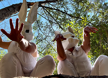 Both dancers are crouching down, looking at the camera from above. They are both dressed in white. Jill Randall is wearing a white bunny mask, and Sima Belmar is wearing a white fox mask